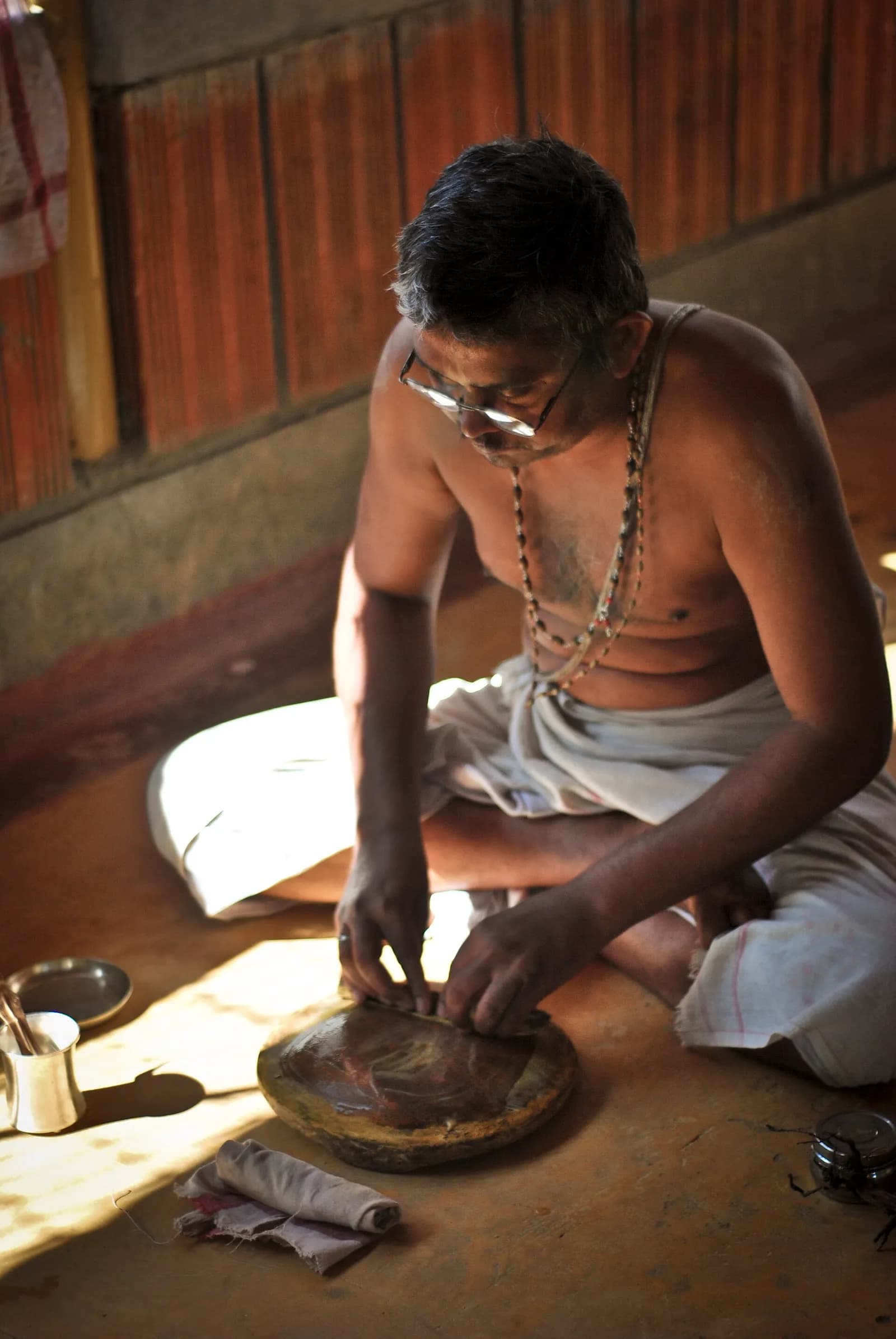 A man sits on the floor grinding a billet of sandalwood on a round stone quern, with vessels for water and paste beside him